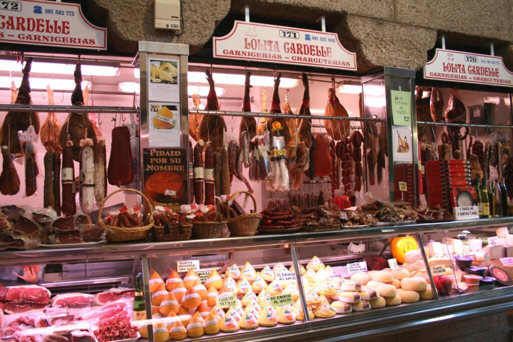 Hams, sausages, and other meats hanging on display above a counter with local cheeses at the Mercado de Abastos