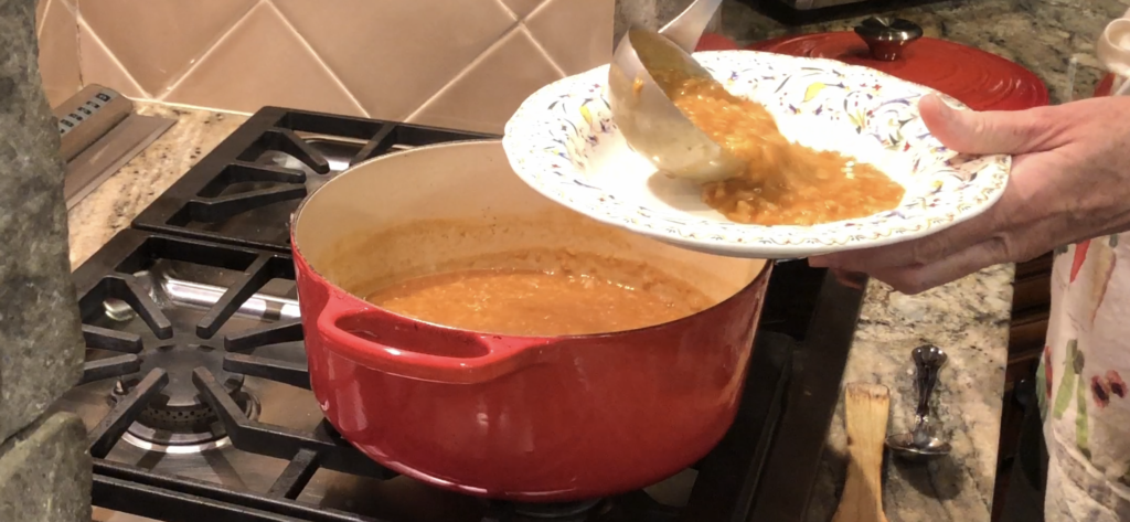 hands ladle red lentil soup from an orange soup pot into a decorative bowl